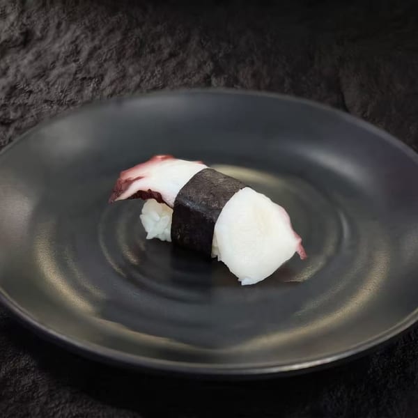 A single piece of octopus nigiri sushi with a dark seaweed wrap on a black plate.
